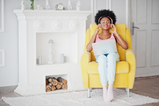 Jovem afro-americana sentada na cadeira amarela enquanto trabalha no laptop e ouve música em fones de ouvido