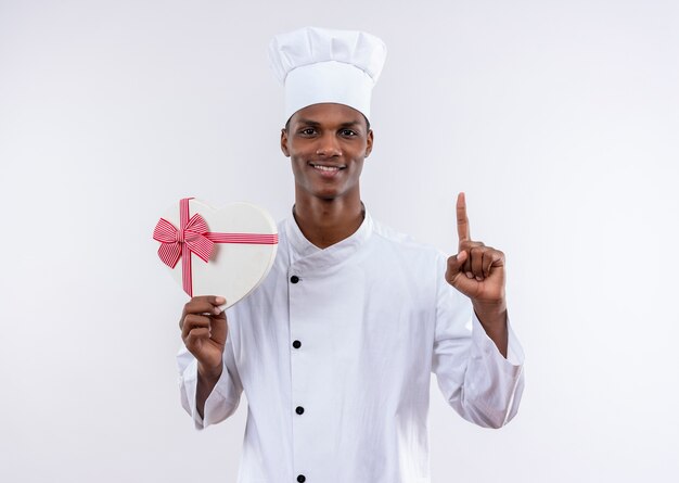Jovem afro-americana sorridente com uniforme de chef segurando uma caixa em forma de coração e pontos isolados no fundo branco com espaço de cópia