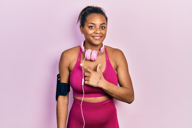 Jovem afro-americana vestindo roupas de ginástica e usando fones de ouvido alegre com um sorriso no rosto apontando com a mão e o dedo para o lado com expressão feliz e natural