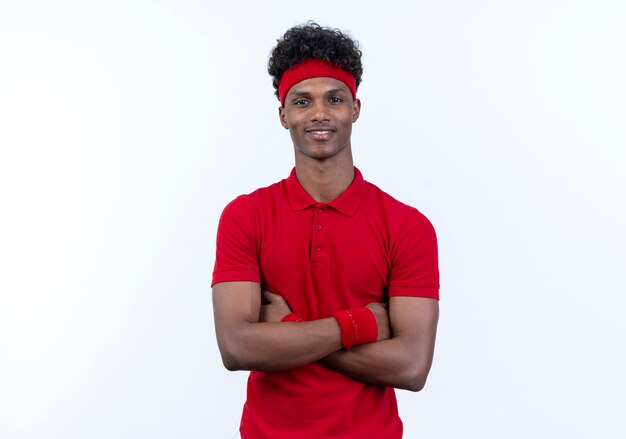 Jovem afro-americano alegre e esportivo usando bandana e pulseira, cruzando as mãos isoladas no fundo branco