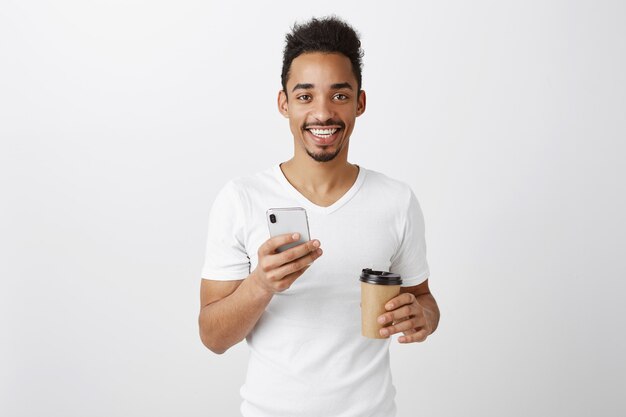 Jovem afro-americano atraente parecendo satisfeito, segurando um celular e bebendo café para viagem