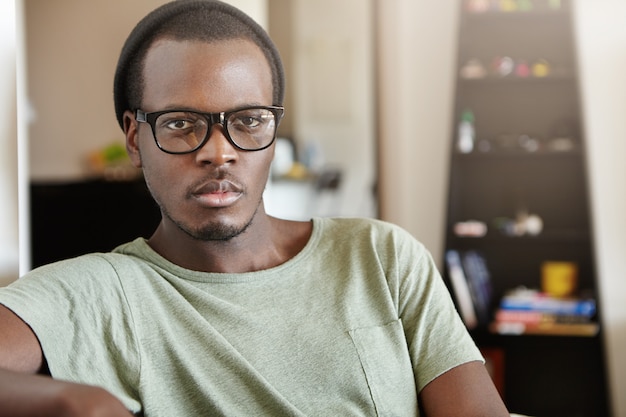 Jovem afro-americano atraente usando óculos escuros na moda e chapéu, passar a manhã de fim de semana em casa, sentado na sala, assistindo as notícias do mundo na tv, tendo um olhar sério