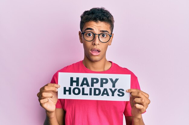 Foto grátis jovem afro-americano bonito segurando mensagem de boas festas com cara de choque, parecendo cético e sarcástico, surpreso com a boca aberta