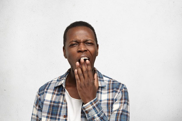 Foto grátis jovem afro-americano cansado ou entediado, cobrindo a boca enquanto boceja, sentindo-se exausto após um dia duro no trabalho. estudante do sexo masculino preto tendo sono chato olhar durante aula de história na faculdade