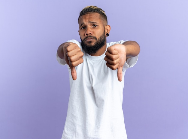 Jovem afro-americano com camiseta branca olhando para a câmera e mostrando os polegares para baixo em pé sobre um fundo azul