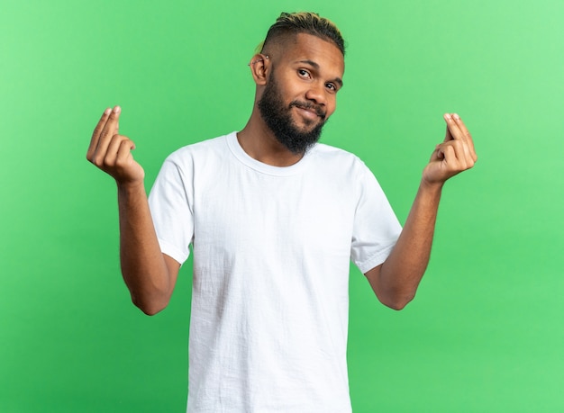 Jovem afro-americano com camiseta branca olhando para a câmera sorrindo, fazendo gesto de dinheiro esfregando os dedos