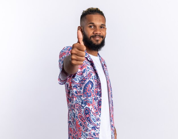 Jovem afro-americano com uma camisa colorida olhando para a câmera, sorrindo alegremente, mostrando os polegares em pé sobre o branco