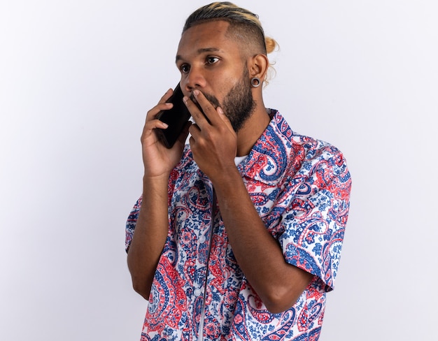 Jovem afro-americano com uma camisa colorida parecendo surpreso ao falar ao telefone celular em pé sobre o branco