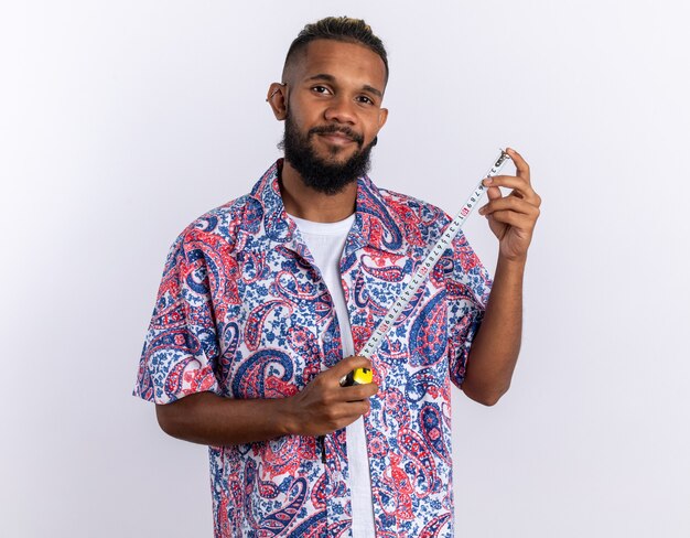 Jovem afro-americano com uma camisa colorida segurando uma fita métrica, olhando para a câmera com um sorriso no rosto feliz e positivo em pé sobre um fundo branco