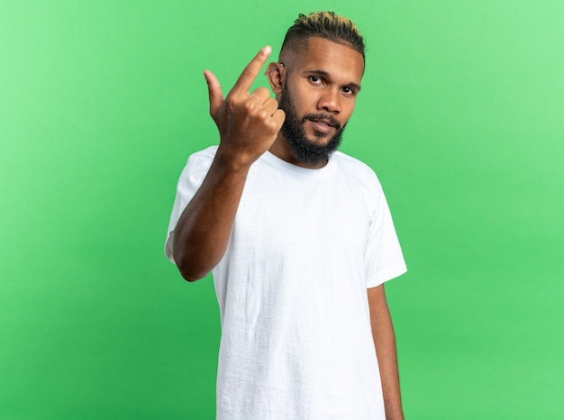 Jovem afro-americano com uma camiseta branca olhando para a câmera com uma cara séria, mostrando o dedo indicador em pé sobre um fundo verde