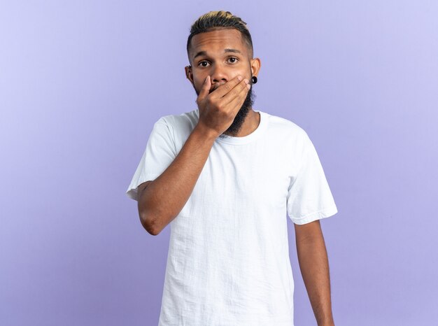 Foto grátis jovem afro-americano com uma camiseta branca olhando para a câmera levando um choque, cobrindo a boca com a mão