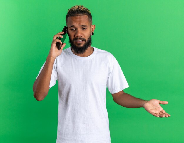 Jovem afro-americano com uma camiseta branca parecendo confuso enquanto fala no celular