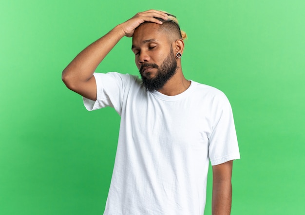 Jovem afro-americano com uma camiseta branca parecendo doente e deprimido com a mão na cabeça em pé sobre um fundo verde
