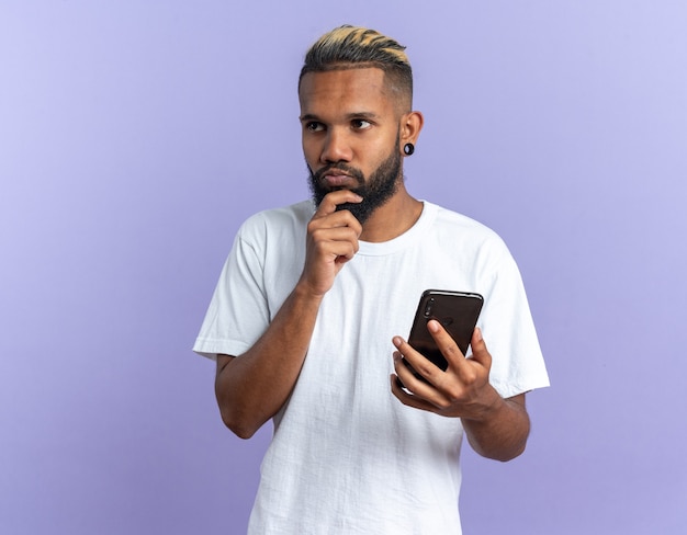 Jovem afro-americano com uma camiseta branca segurando um smartphone e olhando de lado perplexo em pé sobre um fundo azul