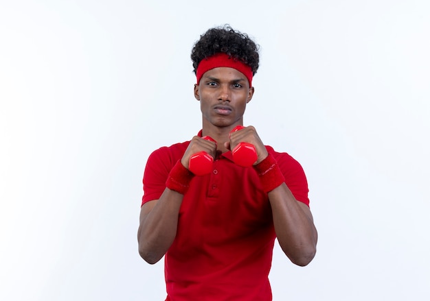 Jovem afro-americano confiante e esportivo, usando bandana e pulseira em pé em pose de luta e segurando halteres isolados no fundo branco