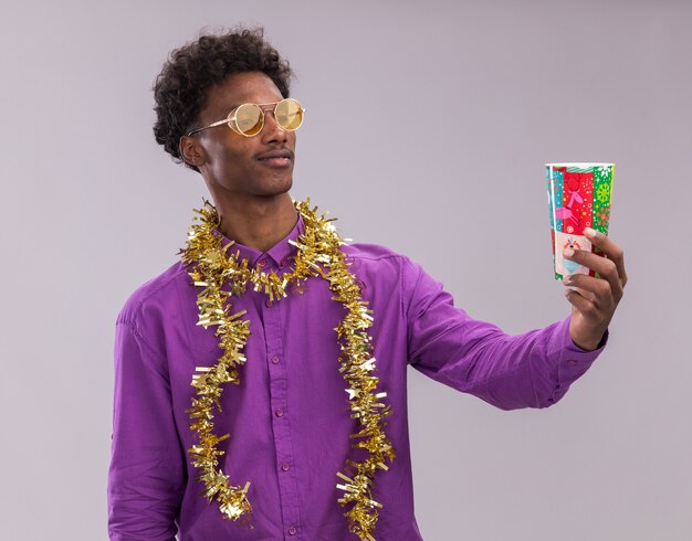 Jovem afro-americano confuso usando óculos com guirlanda de ouropel no pescoço, esticando o copo de plástico de Natal, olhando para ele isolado no fundo branco