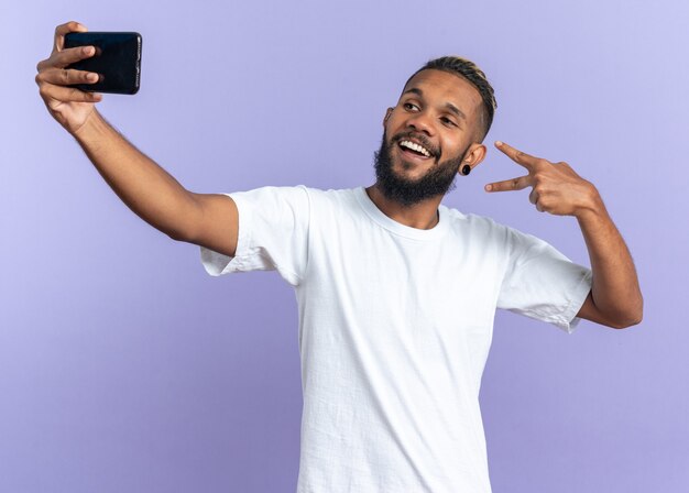 Jovem afro-americano de camiseta branca fazendo selfie usando smartphone, sorrindo alegremente, mostrando o sinal V em pé sobre fundo azul