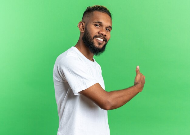 Jovem afro-americano de camiseta branca olhando para a câmera, sorrindo alegremente, apontando para trás com o dedo em pé sobre um fundo verde