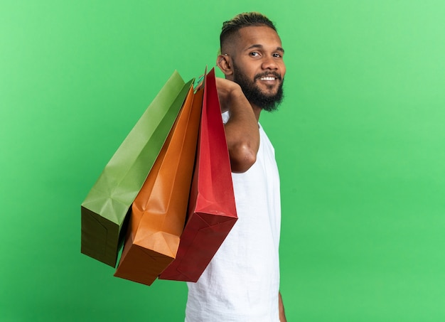 Jovem afro-americano de camiseta branca segurando sacolas de papel, olhando para a câmera, sorrindo alegremente feliz e positivo