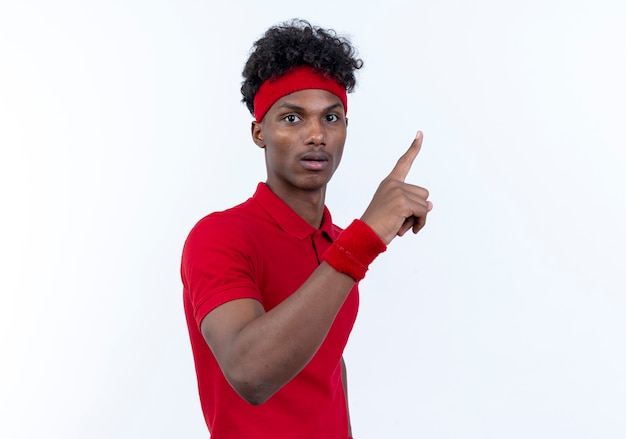 Jovem afro-americano e esportivo impressionado usando a fita para a cabeça e as pontas da pulseira isoladas no fundo branco com espaço de cópia