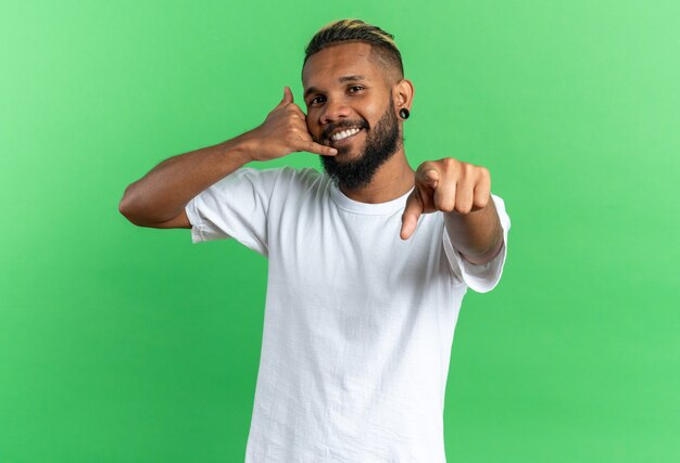 Jovem afro-americano em uma camiseta branca apontando com o dedo indicador para a câmera, fazendo um gesto de me chamar de sorriso amigável em pé sobre um fundo verde