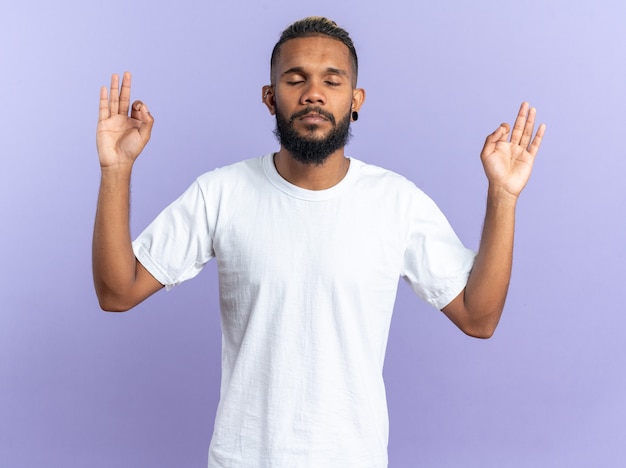 Jovem afro-americano em uma camiseta branca tentando relaxar fazendo gestos de meditação com os dedos e os olhos fechados