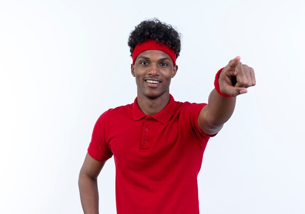 Jovem afro-americano esportivo sorridente usando bandana e pulseira apontando para a câmera isolada no branco