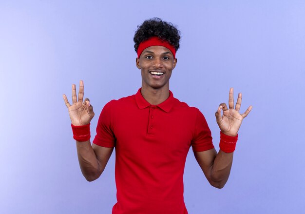 Jovem afro-americano esportivo sorridente usando bandana e pulseira, mostrando gesto ok isolado em azul