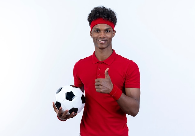 Jovem afro-americano esportivo sorridente usando bandana e pulseira, segurando uma bola com o polegar isolado no branco