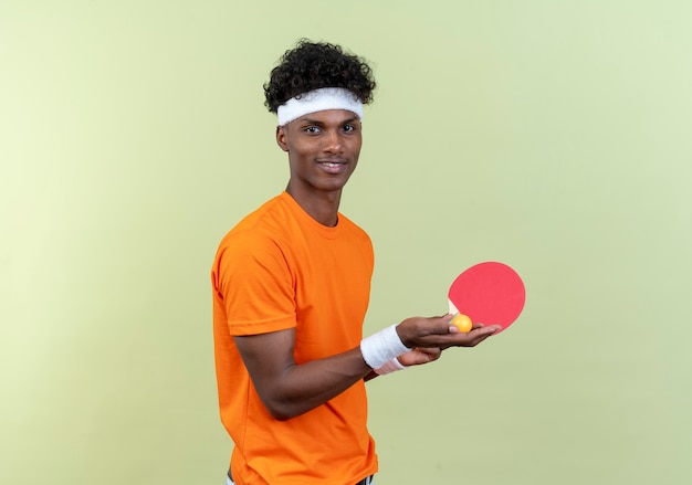 Jovem afro-americano feliz e esportivo, usando bandana e pulseira, segurando uma raquete de pingue-pongue com uma bola isolada no fundo verde