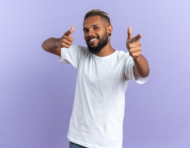 Jovem afro-americano feliz e satisfeito com uma camiseta branca apontando com o dedo indicador para a câmera, sorrindo alegremente em pé sobre um fundo azul