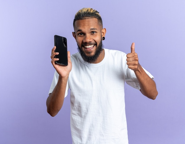 Jovem afro-americano feliz em uma camiseta branca segurando um smartphone, olhando para a câmera mostrando os polegares sorrindo alegremente