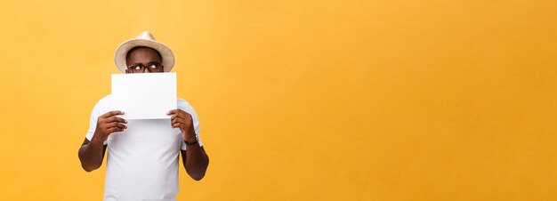 Jovem afro-americano feliz escondido atrás de um papel em branco isolado em fundo amarelo