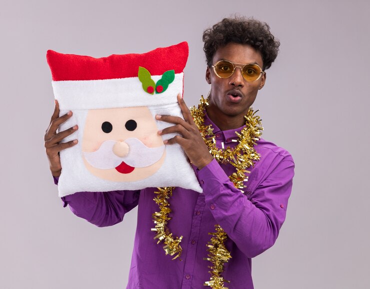 Foto grátis jovem afro-americano impressionado usando óculos com guirlanda de ouropel no pescoço, segurando a almofada de papai noel, olhando para a câmera isolada no fundo branco