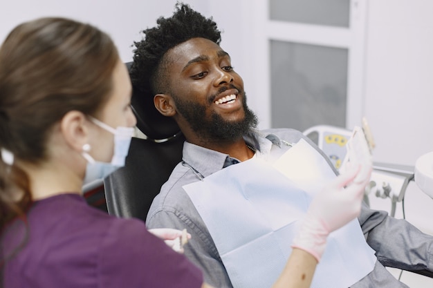 Jovem afro-americano. Indivíduo em visita ao consultório dentário para prevenção da cavidade oral. Homem e médico famoso durante o exame de dentes.