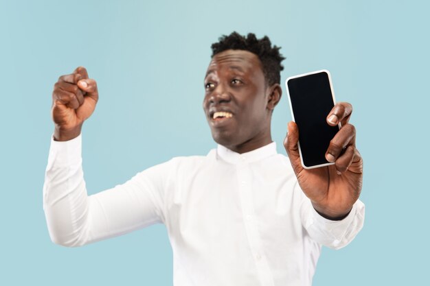 Jovem afro-americano posando com smartphone isolado em um fundo azul do estúdio, expressão facial. Belo retrato masculino de meio comprimento. Conceito de emoções humanas, expressão facial.