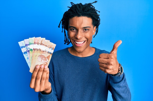 Jovem afro-americano segurando dólares canadenses sorrindo feliz e positivo polegar para cima fazendo excelente e sinal de aprovação