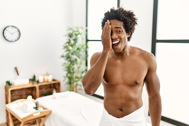 Jovem afro-americano sem camisa vestindo toalha em pé no centro de beleza cobrindo um olho com a mão sorriso confiante no rosto e emoção surpresa