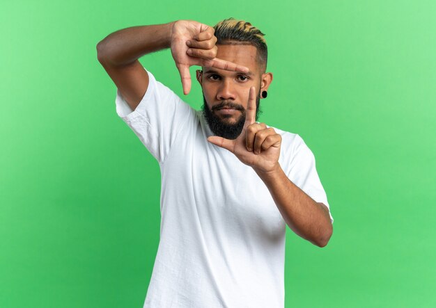 Jovem afro-americano sério com camiseta branca fazendo moldura com os dedos olhando por entre os dedos