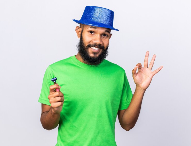 Foto grátis jovem afro-americano sorridente com chapéu de festa, mostrando o gesto de ok, segurando o apito de festa