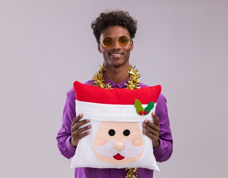 Foto grátis jovem afro-americano sorridente, usando óculos com guirlanda de ouropel no pescoço, segurando a almofada de papai noel, olhando para a câmera, isolada no fundo branco