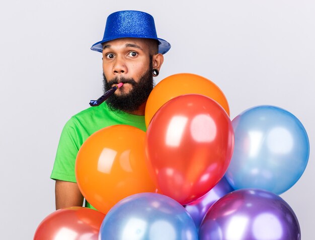 Jovem afro-americano surpreso com um chapéu de festa segurando balões e soprando um apito de festa
