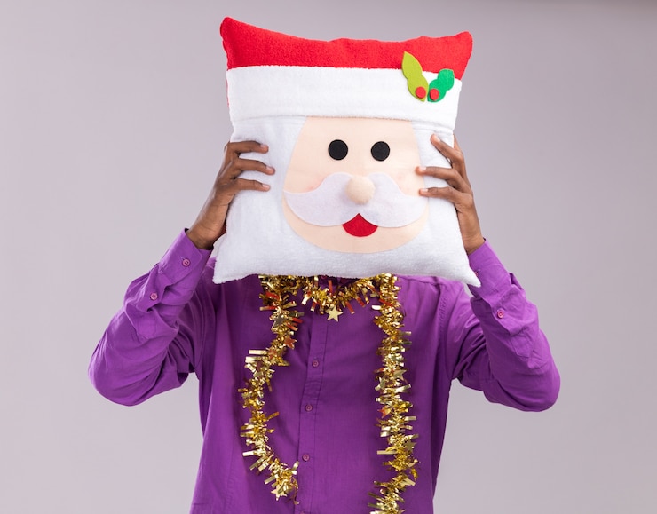 Foto grátis jovem afro-americano usando óculos com guirlanda de ouropel no pescoço, segurando a almofada de papai noel na frente do rosto, isolado no fundo branco