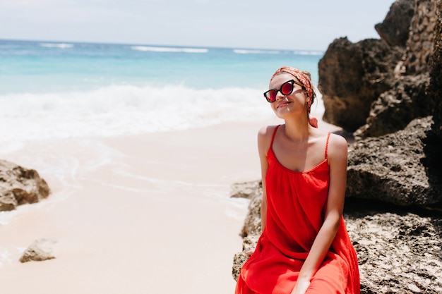 Jovem agradável tomando banho de sol na praia selvagem. Menina europeia sorridente em traje vermelho sentada na rocha com o oceano