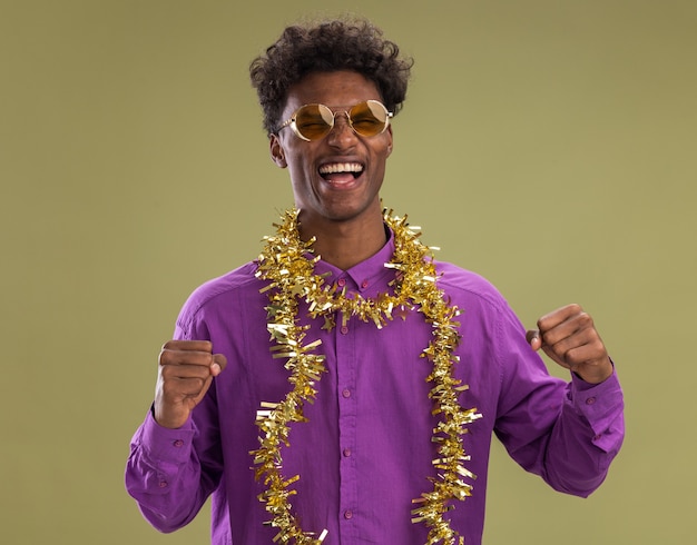 Foto grátis jovem alegre afro-americano de óculos com guirlanda de ouropel no pescoço, olhando para a câmera, fazendo gesto de sim isolado em fundo verde oliva