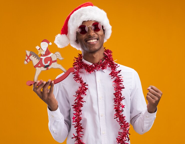 Foto grátis jovem alegre afro-americano usando chapéu de papai noel e óculos com guirlanda de ouropel em volta do pescoço segurando o papai noel na estatueta de cavalo de balanço, olhando para a câmera, fazendo gesto de sim isolado em fundo laranja