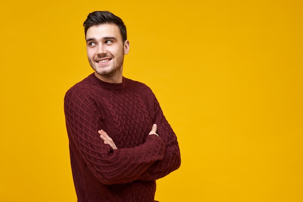 Jovem alegre atraente vestindo blusa de malha, estando de bom humor, olhando para longe com um sorriso feliz e confiante, segurando os braços cruzados sobre o peito. Cara bonito posando de jumper isolado