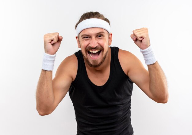 Jovem alegre, bonito e desportivo usando bandana e pulseiras levantando os punhos isolados na parede branca