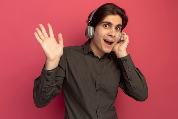 Jovem alegre bonito vestindo uma camiseta preta com fones de ouvido mostrando um gesto de olá isolado na parede rosa