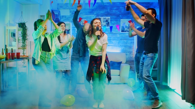 Jovem alegre cantando para seus amigos na festa em uma sala com luzes de néon e fumaça.